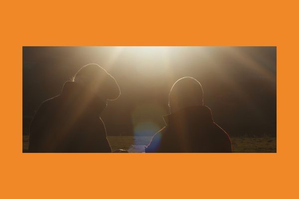 Two men in winter coats stand in a field with the sun beaming down on them.