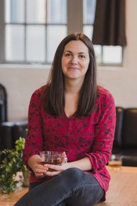 A woman with brown hair smiling. She is wearing a red top with dark spots and black jeans.