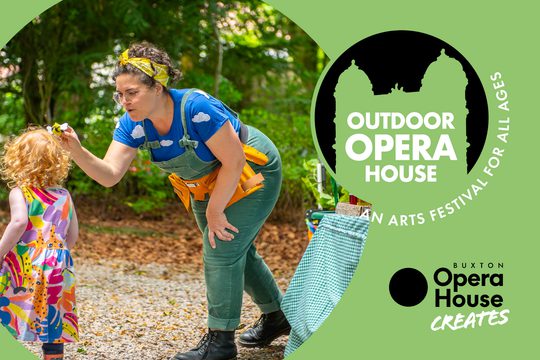 A woman in green dungarees and a blue top with clouds on it holds a small fuzzy bee up to a young girl.