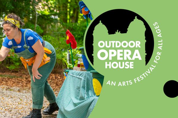A woman in green dungarees and a blue top with clouds on it holds a small fuzzy bee up to a young girl.