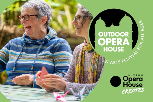 Two women wearing glasses laughing and doing craft on a picnic bench. Outdoor Opera House logo and Buxton Opera House Creates logo.