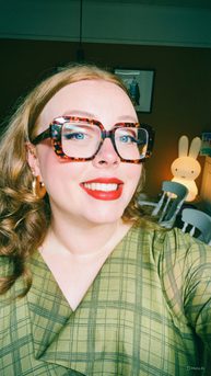 A woman with ginger hair and red lipstick smiling. She is wearing large glasses and a green dress.