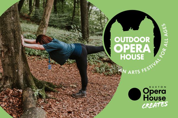 A woman in a green top doing yoga in a forest. Her pose is supported by a tree trunk and she is standing on one leg.