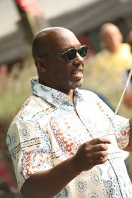 A man wearing sunglasses and a colourful shirt holding a conducting baton.