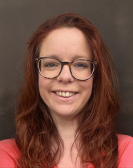 A woman smiling. She has red hair and is wearing glasses and a bright pink top.
