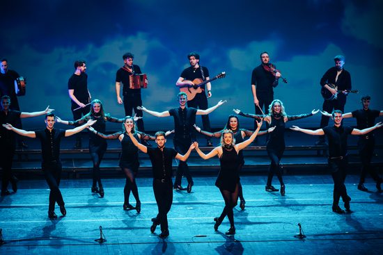 A group of dancers dressed in black. They are all smiling, with one leg crossed in front of the other, and their arms stretched out wide.