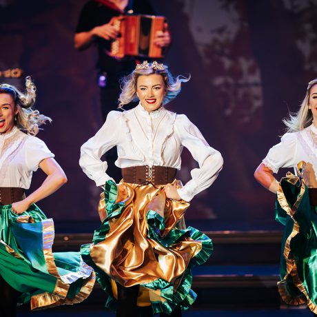 Three women dance in white shirts, thick brown belts, and green and gold skirts.