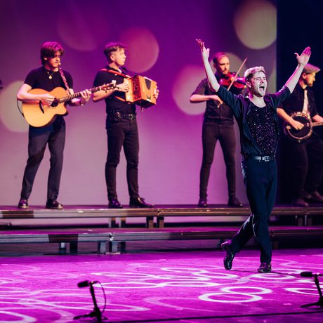 Five musicians play instruments at the back of the stage, while a dancer performs in front of them.