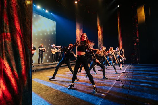A group of dancers wearing black on a stage. They are all facing the same direction, with their arms out on either side of them.