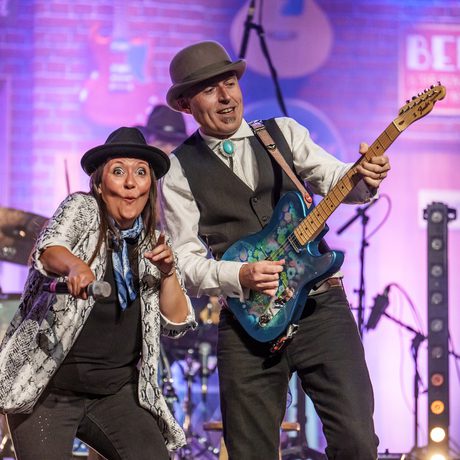 A man and a woman both wearing hats having a laugh together on stage.