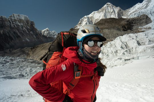 A woman in a red coat and helmet standing on a snowy mountain, carrying a large backpack.