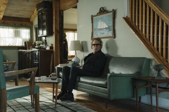 An older white man with curly hair wears a dark suit and sunglasses. He sits in a living room in a green leather sofa.