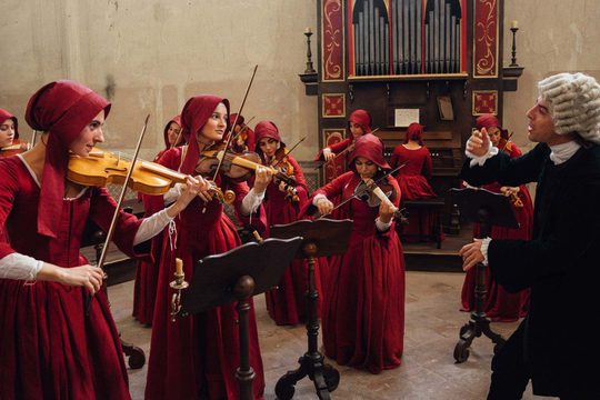 A group of young girls in red dresses and bonnets play violins. They are conducted by a man in a black suit wearing a grey wig.