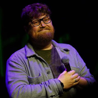 A man with curly hair and a beard holds a microphone and smiles.