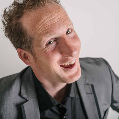 A man in a grey suit smiling widely and leaning towards the camera.