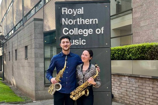 One man and one woman standing holding saxophones. The sign behind them reads Royal Northern College of Music.