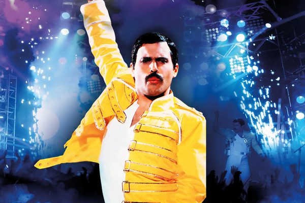 Freddie Mercury in a yellow jacket. He has one arm in the air.