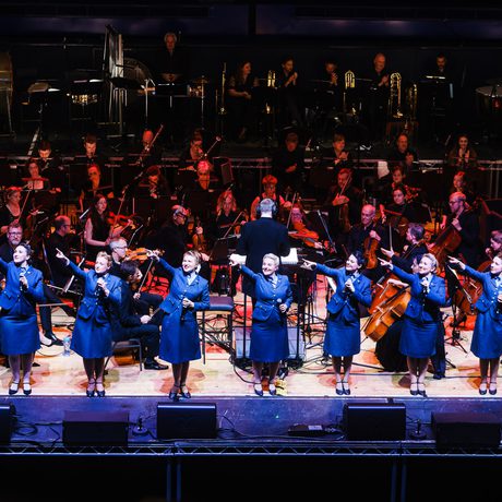 A group of women in blue skirts and jackets. They are standing in a line and pointing upwards.
