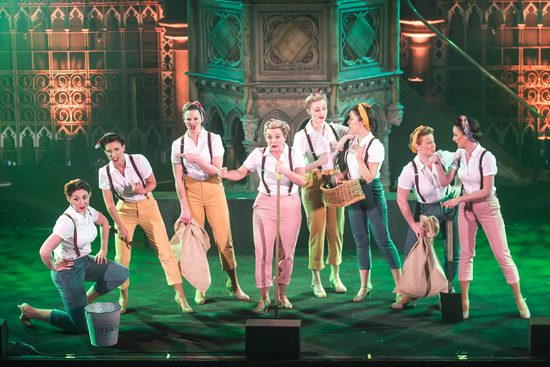 A group of women in white shirts, colourful trousers, and suspenders, surrounded by green lighting.