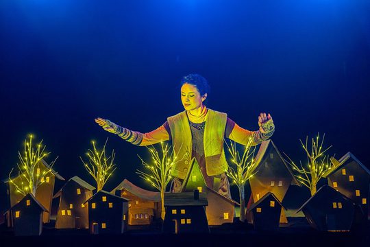 A woman in a green vest stands over a small wooden model village, lit up by small model trees.