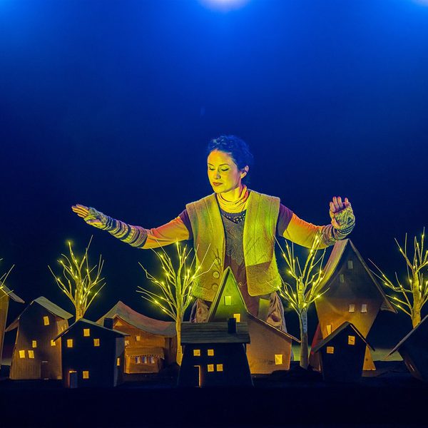 A woman in a green vest stands over a small wooden model village, lit up by small model trees.