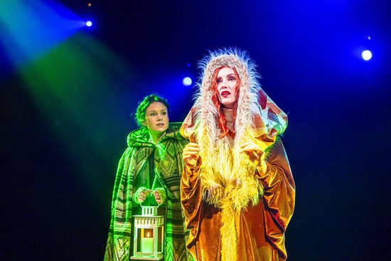 A woman in a green cloak carrying a lantern speaks to another woman in a large yellow cloak with fur around the hood.