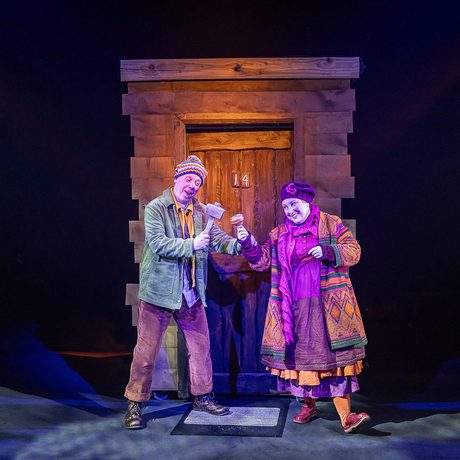 A man in a green jacket and brown trousers stands with a woman wearing a multicoloured cardigan and purple hat outside a wooden door with the number 14 on it.