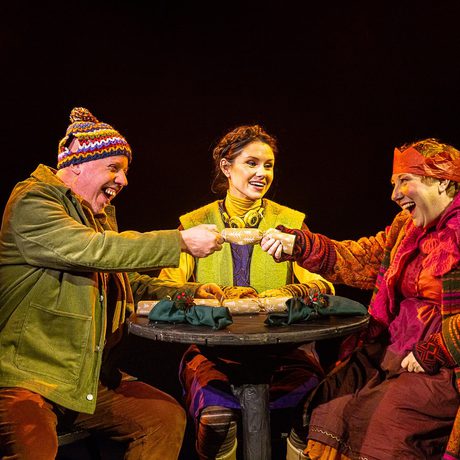 Three people in colourful clothes and woollen hats sit at a wooden table and pull Christmas crackers.