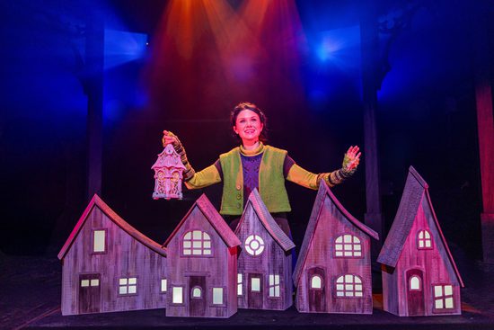 A woman in a green knitted vest holds a lantern over a wooden model village.