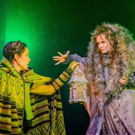 A woman in a green cloak holds a lantern up to another woman in a grey cloak with a large fur hood. They are lit by green lighting.