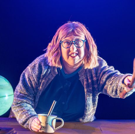 A woman with shoulder-length blonde hair and blue glasses stands behind a desk with a globe on it and waves a stick forwards.