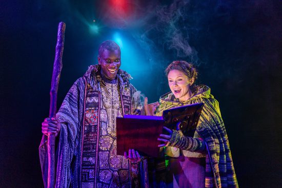 A man in a patterned cloak holding a wooden staff. He is looking at a large book with a woman wearing a green cloak.