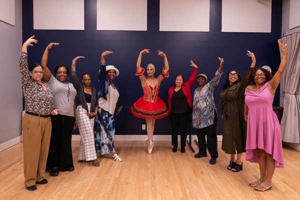 A group of women with one arm above their head. In the centre is a ballerina with both arms above their head, wearing red and standing en pointe.