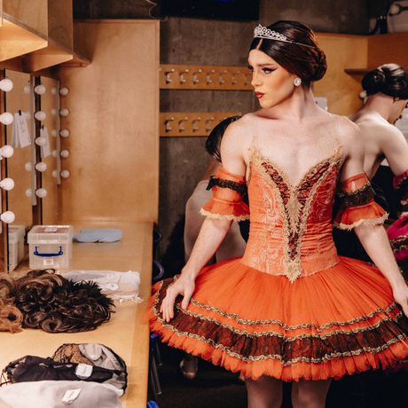 A male ballet dancer wearing a brunette wig and orange tutu looks in the mirror.
