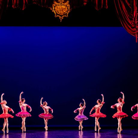 A line of male ballet dancers wearing pink and purple tutus. They are all en pointe and have one arm in the air. The stage is surrounded by red curtains.