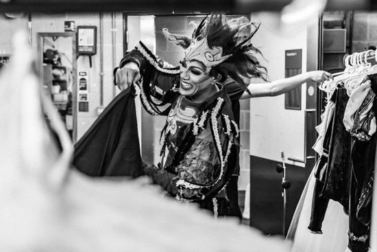 Black and white image of a male dancer in heavy make up and an extravagant costume and headdress smiles backstage.