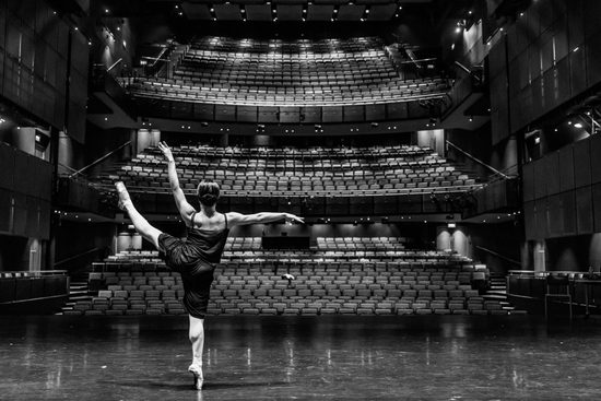 Black and white image of a ballet dancer en pointe from behind. They are practicing on a stage and the seats in the theatre are empty.