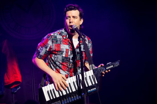 A man in a red and white shirt with varying patterns in it sings into a microphone and plays a piano that he wears like a guitar.