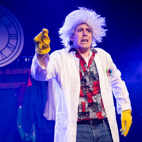 A man in a white lab coat, yellow rubber gloves, and a wig of long white hair.