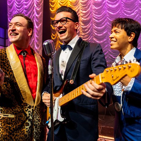 Three men are smiling widely. Two are holding microphones and one is playing the guitar.