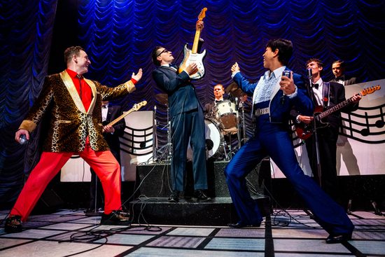 A man in a blue suit plays the guitar while holding it in the air. He has a band and backing singers around him who are watching him.
