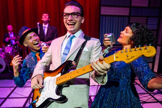 A man in a cream suit and blue tie is smiling and playing the guitar. A man and a woman both dressed in blue are singing on either side of him.