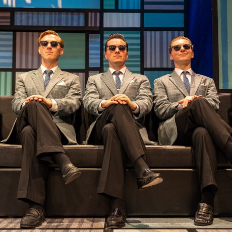 Three men wearing identical grey suits and sunglasses sit on a black sofa with their legs crossed and their hands on their knee.