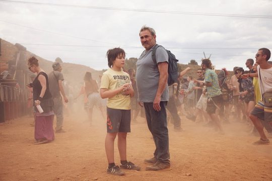 A man and his young son stand with a large group of people. There is sand and dust everywhere.