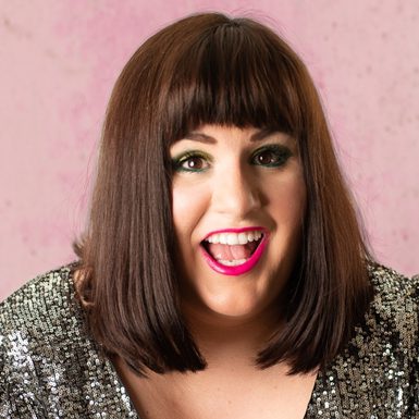 A woman with shoulder length brunette hair and a blunt fringe. She is wearing a sparkly top and bright pink lipstick.