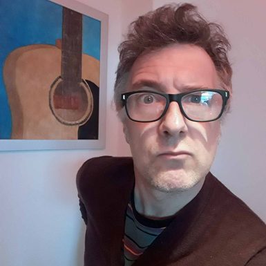 A man with curly hair and glasses stands next to a painting of a guitar.