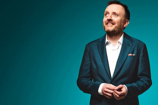 Chris in a suit fixes his cuff links in front of a turquoise background
