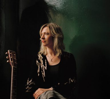A blonde woman sat in front of a dark green background with a guitar next to her.
