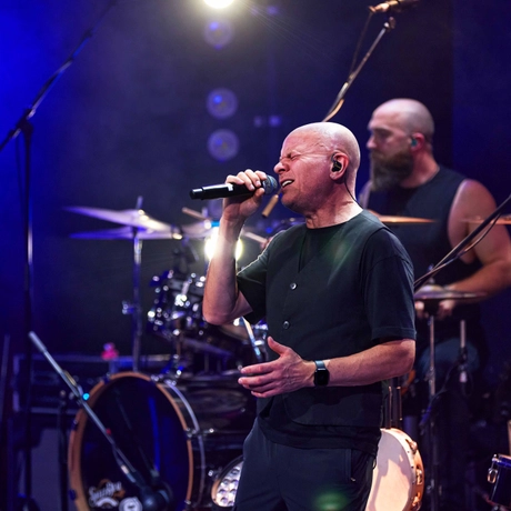 A man in a black t-shirt holds a microphone and sings. His eyes are closed.