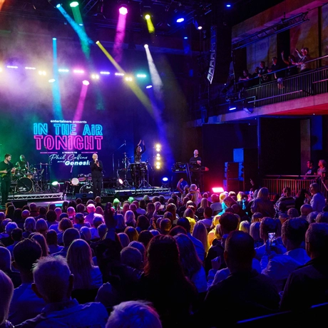 A man singing on stage, with a band playing, and large crowd watching him. There are spotlights of various different colours covering the stage and the audience.